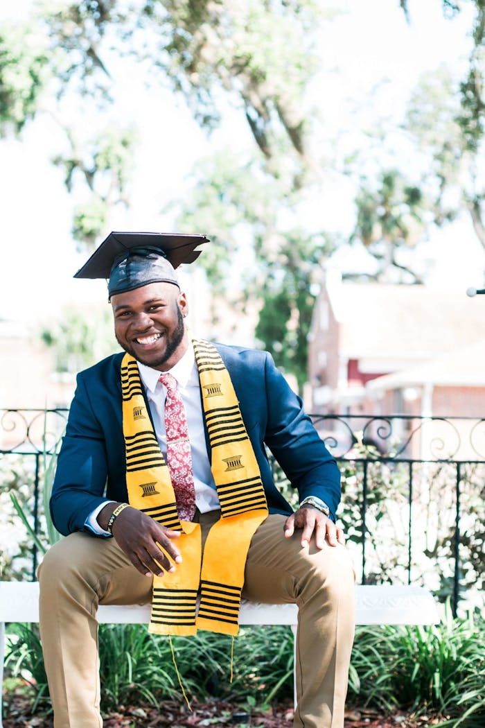 gallery-02 Smiling graduate sitting outside wearing a cap and gown with stole.