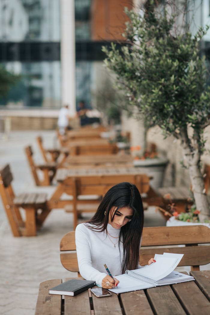 Young focused ethnic female manager taking notes in copybook at wooden table in street cafeteria