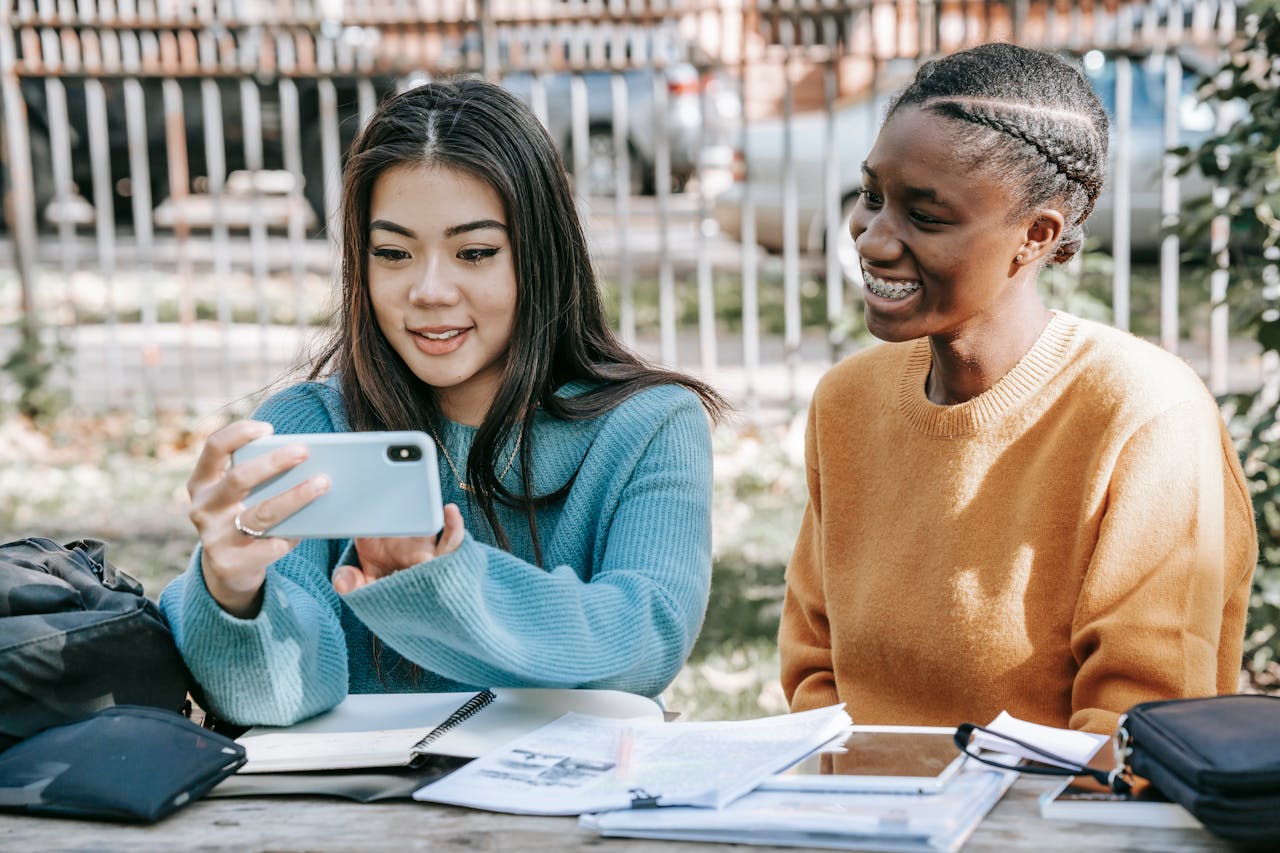 Two young women studying outdoors with a smartphone, smiling and enjoying a casual moment.