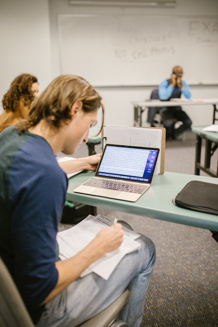College student focused on exam preparation in a classroom setting, using a laptop and notes.