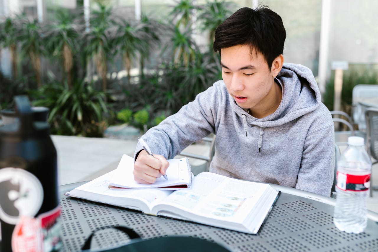 our-experience Asian student in gray hoodie writing in notebook outdoors, studying for university courses.