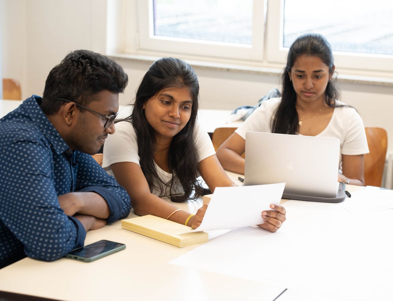 A group of students working together indoors, focused on documents and a laptop.