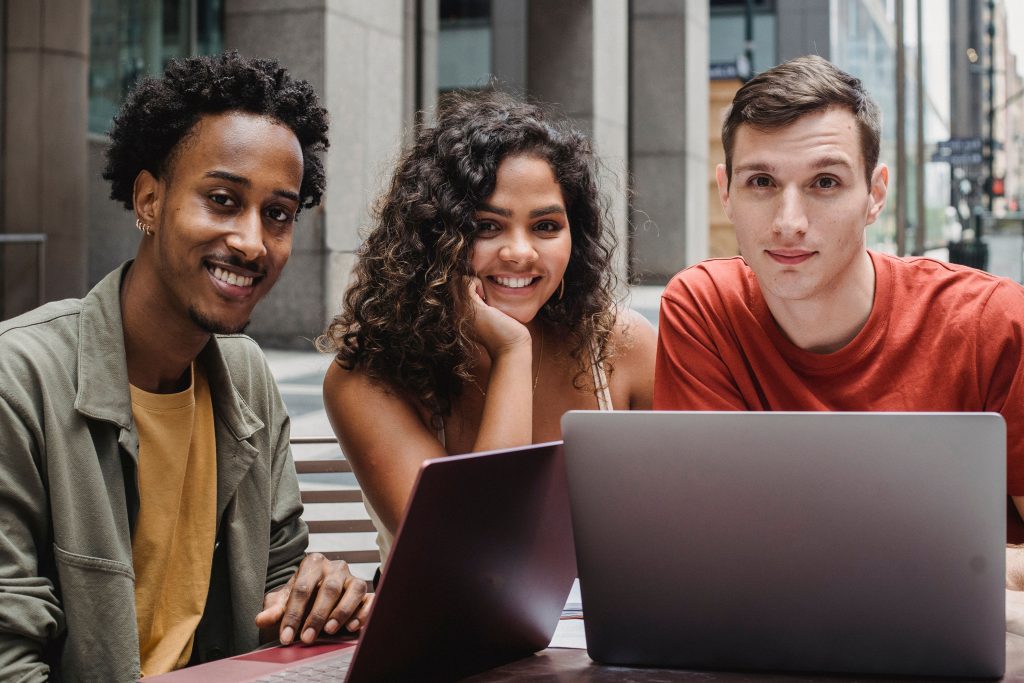 Young multiethnic friends in casual outfit using netbook while sitting at table in city street and looking at camera