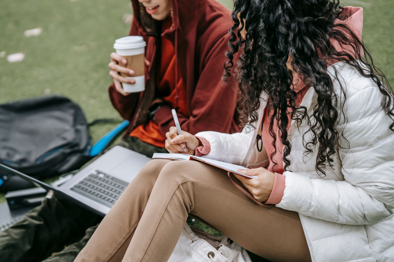 project-02-a Side view of unrecognizable classmates with takeaway coffee browsing laptop and taking notes in notebook while preparing for classes on grassy ground
