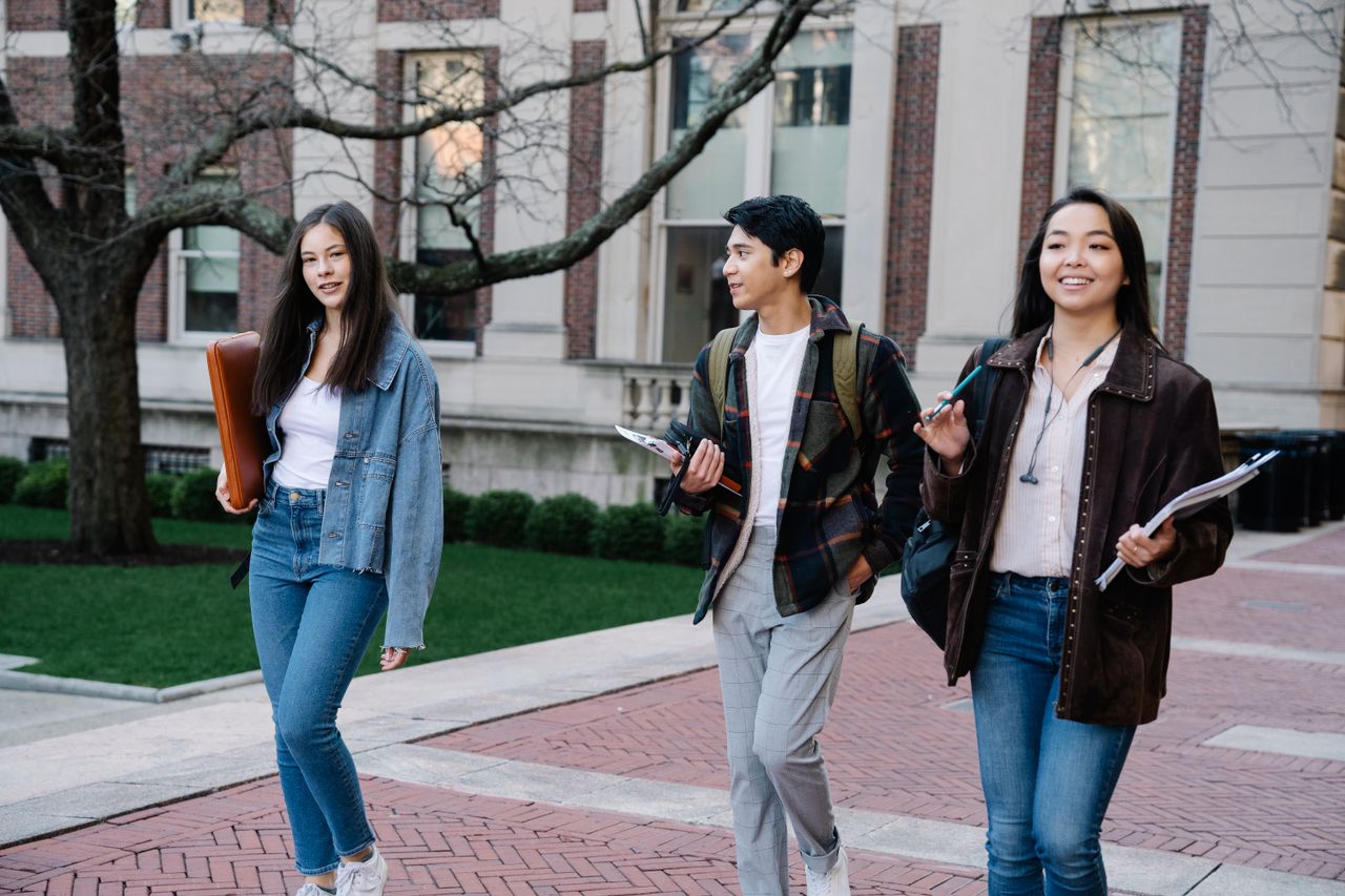Group of college students walking across a campus sidewalk, carrying books and backpacks.
