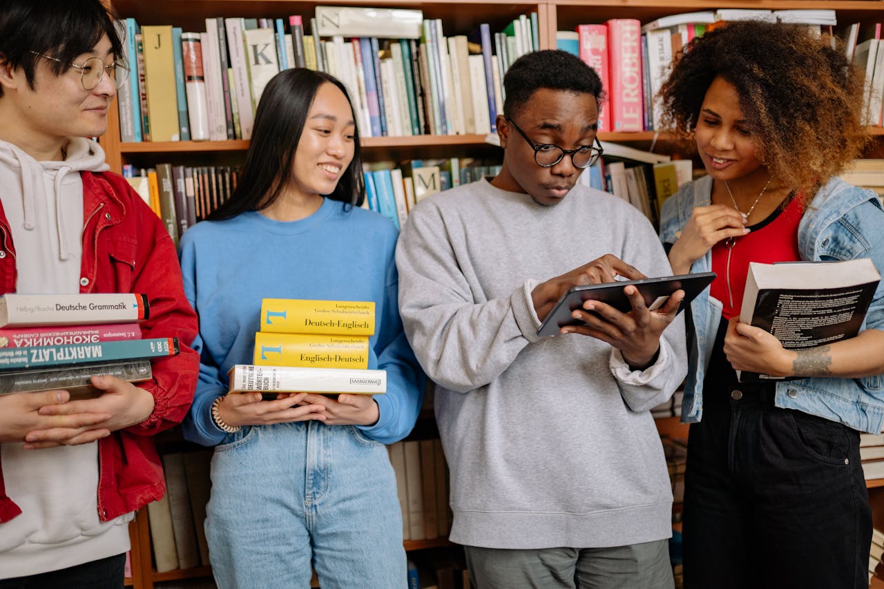 A group of diverse students study together in a library, using books and a tablet for learning.