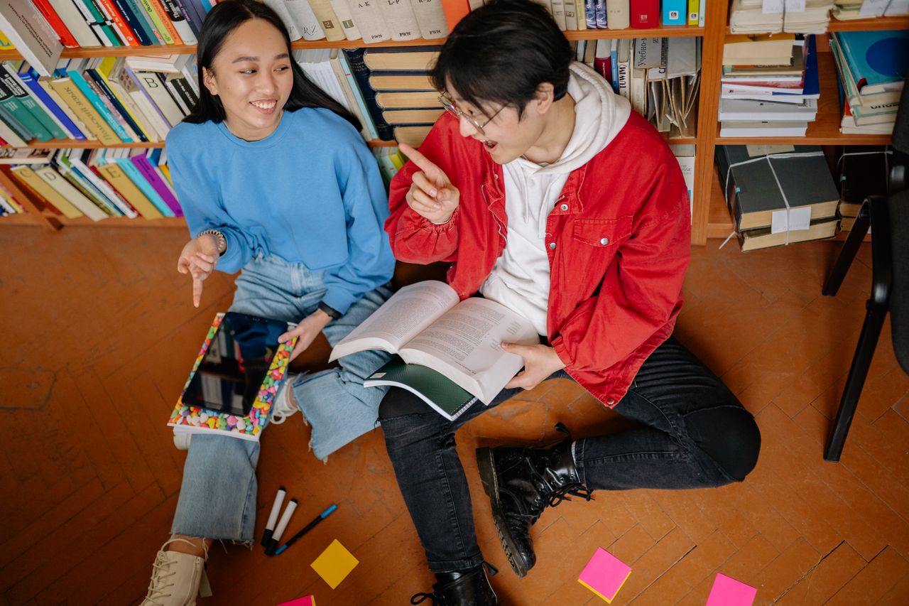 project-03-a Two students laughing and discussing study materials while sitting in a library.