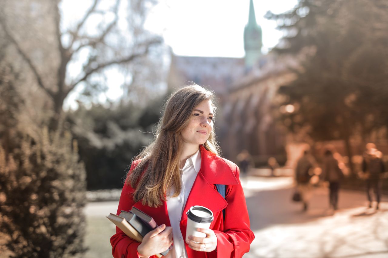 featured-images-03 Smiling woman in red coat with coffee and books, enjoying leisure time outdoors on a university campus.