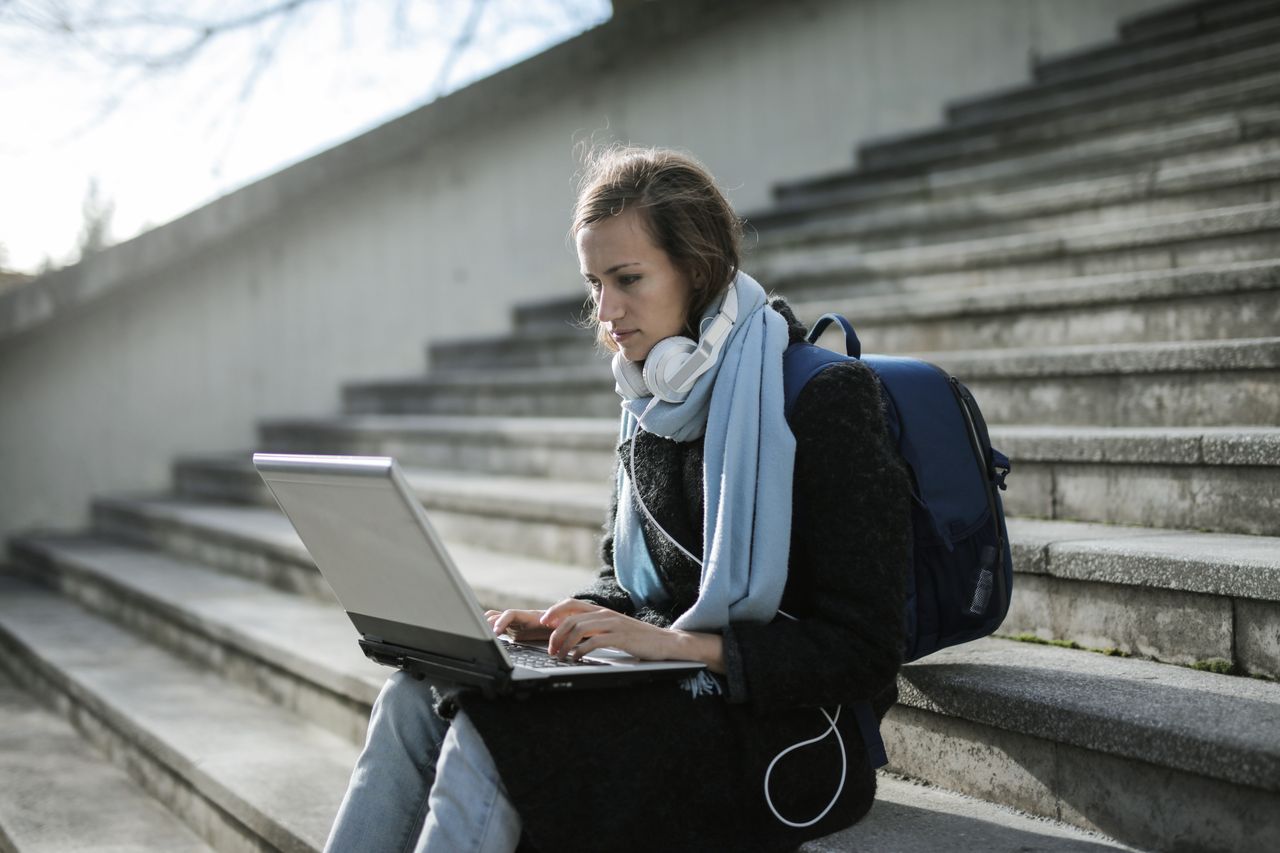 services-04 Woman using laptop on outdoor stairs, studying online with headphones and backpack.