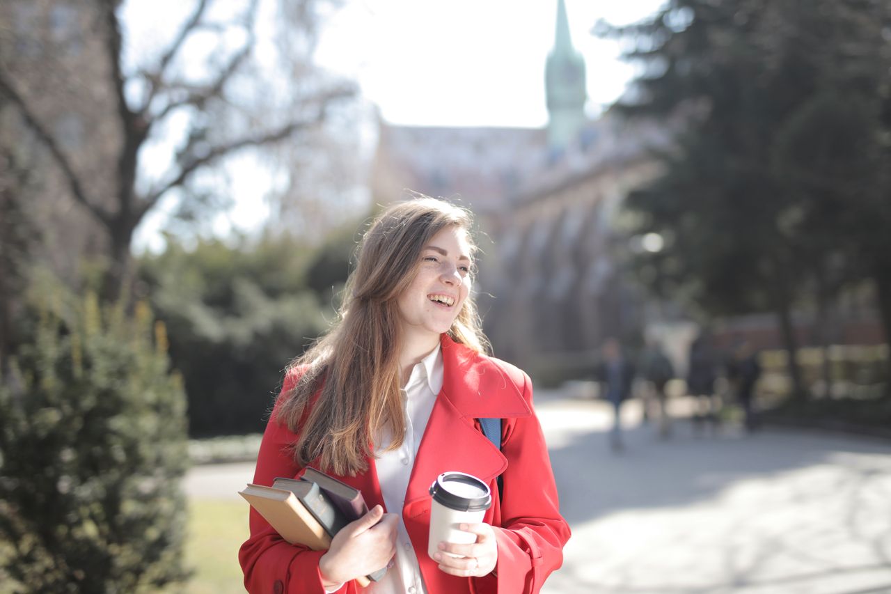featured-images-05 Cheerful young woman in elegant wear with pile of books and hot drink to go standing on lawn of university campus and looking away