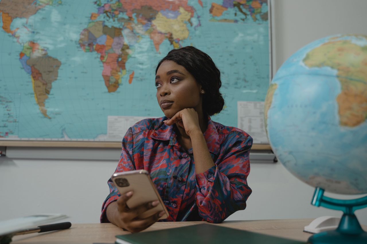 gallery-03 A teacher in a classroom, pondering with a smartphone. Educational setting with a globe and map.