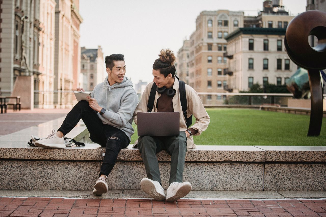 behind-the-scene-05 Full body of young multiracial male friends in casual clothes sitting on stone border in park and working on netbook while making notes in notepad in daytime