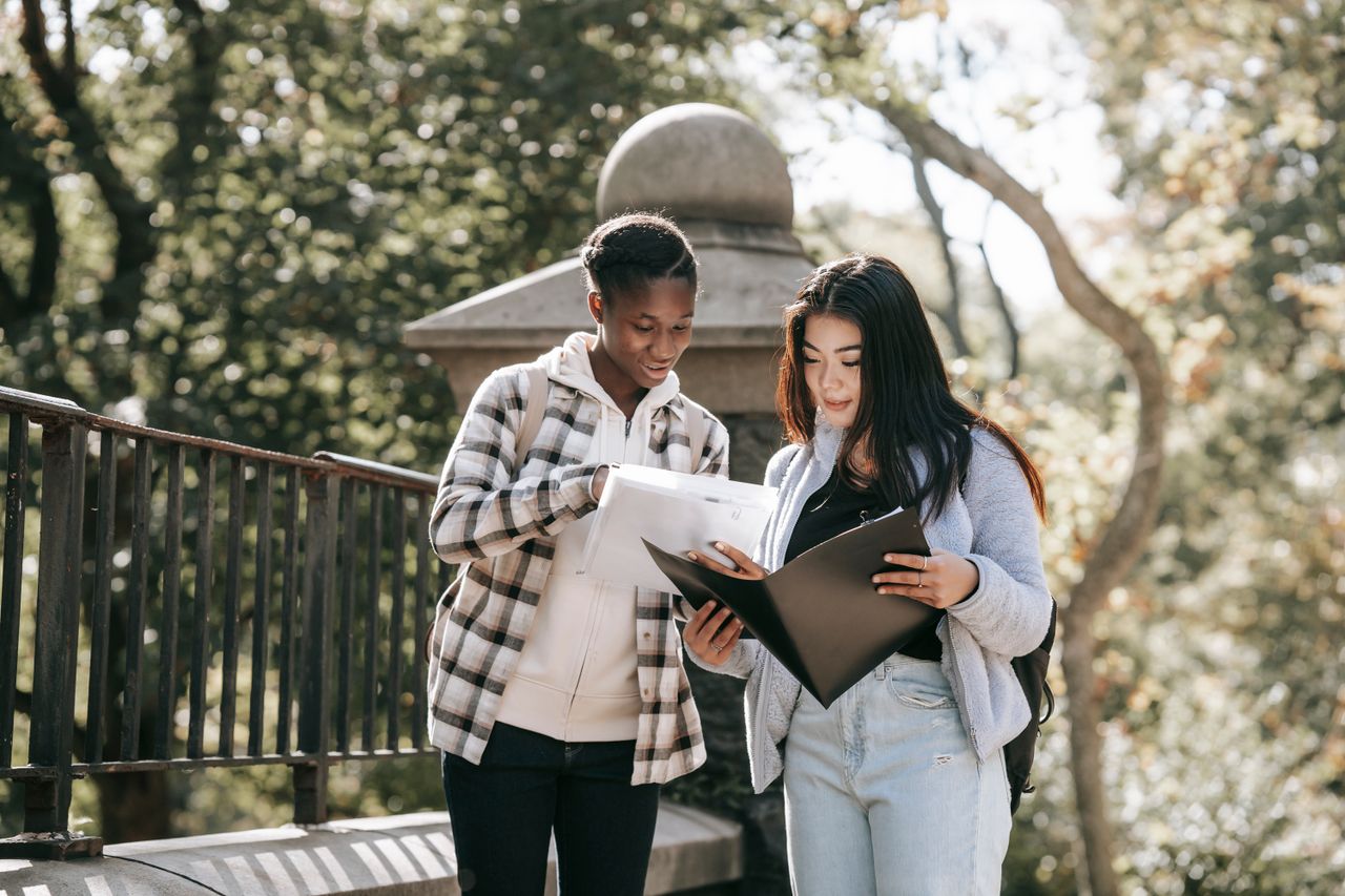 featured-images-02 Young attentive multiracial girlfriends in casual wear talking while watching folders and studying in city on sunny day in back lit