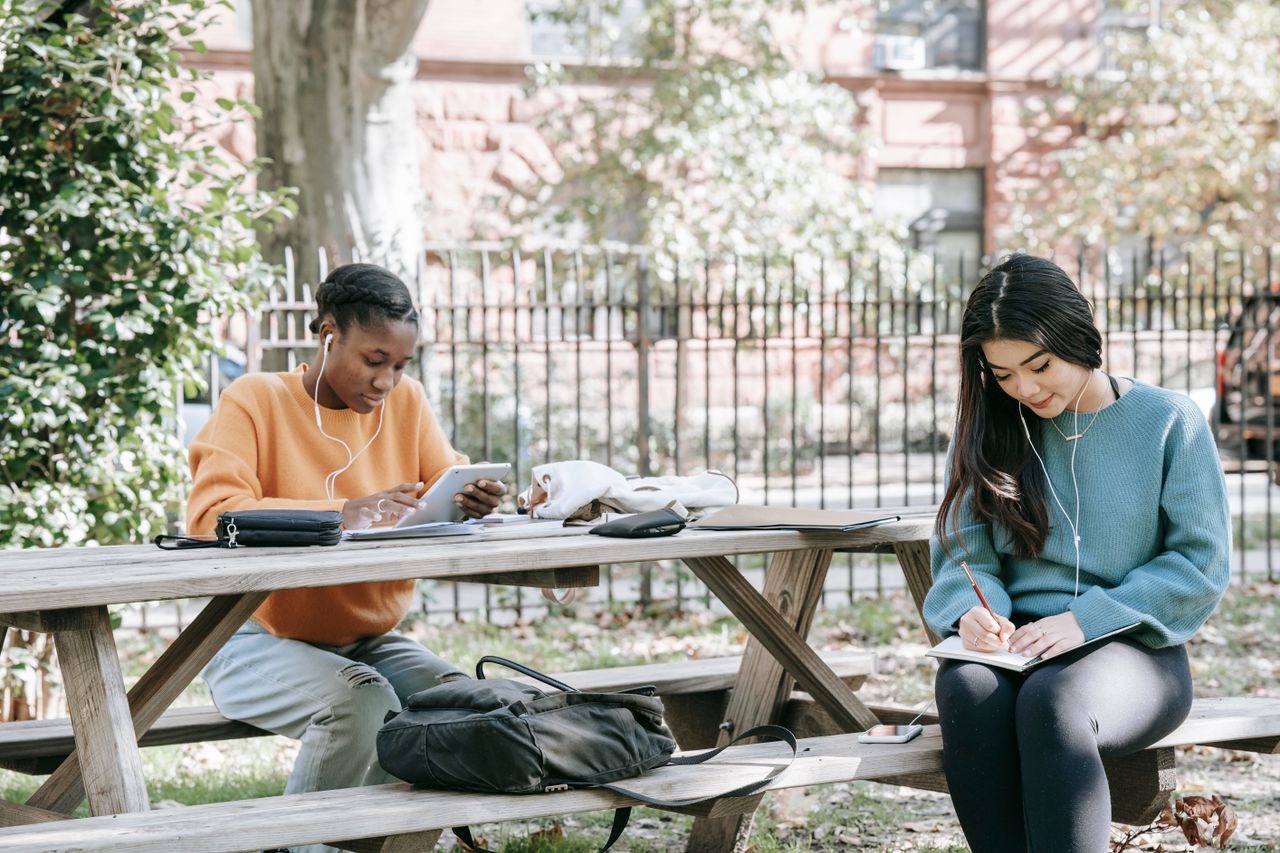why-choose-us-02 Positive young multiracial female students sitting at table in city park and preparing for exams together while browsing tablet and taking notes in copybook