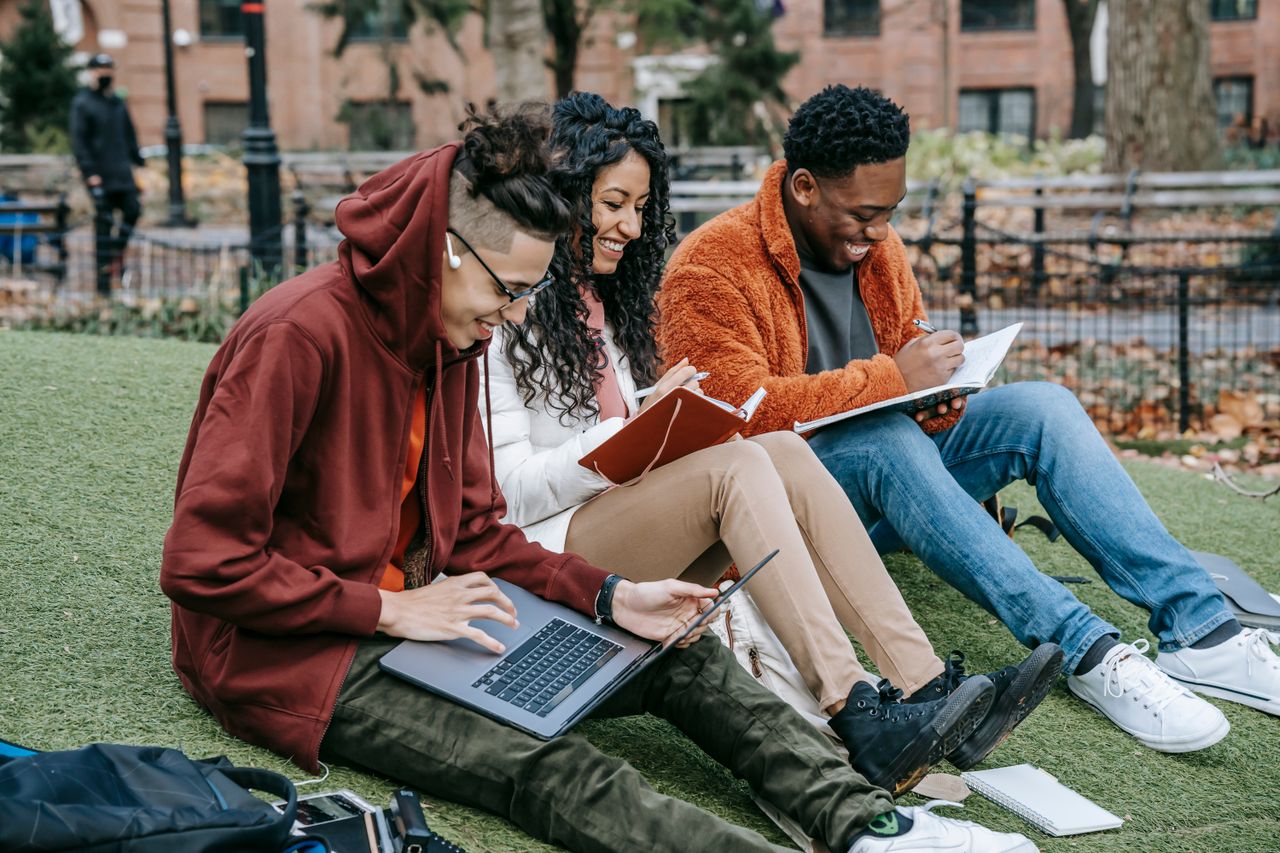featured-images-04 Happy multiracial group of students in casual clothes sitting together on grass and studying with laptop and books