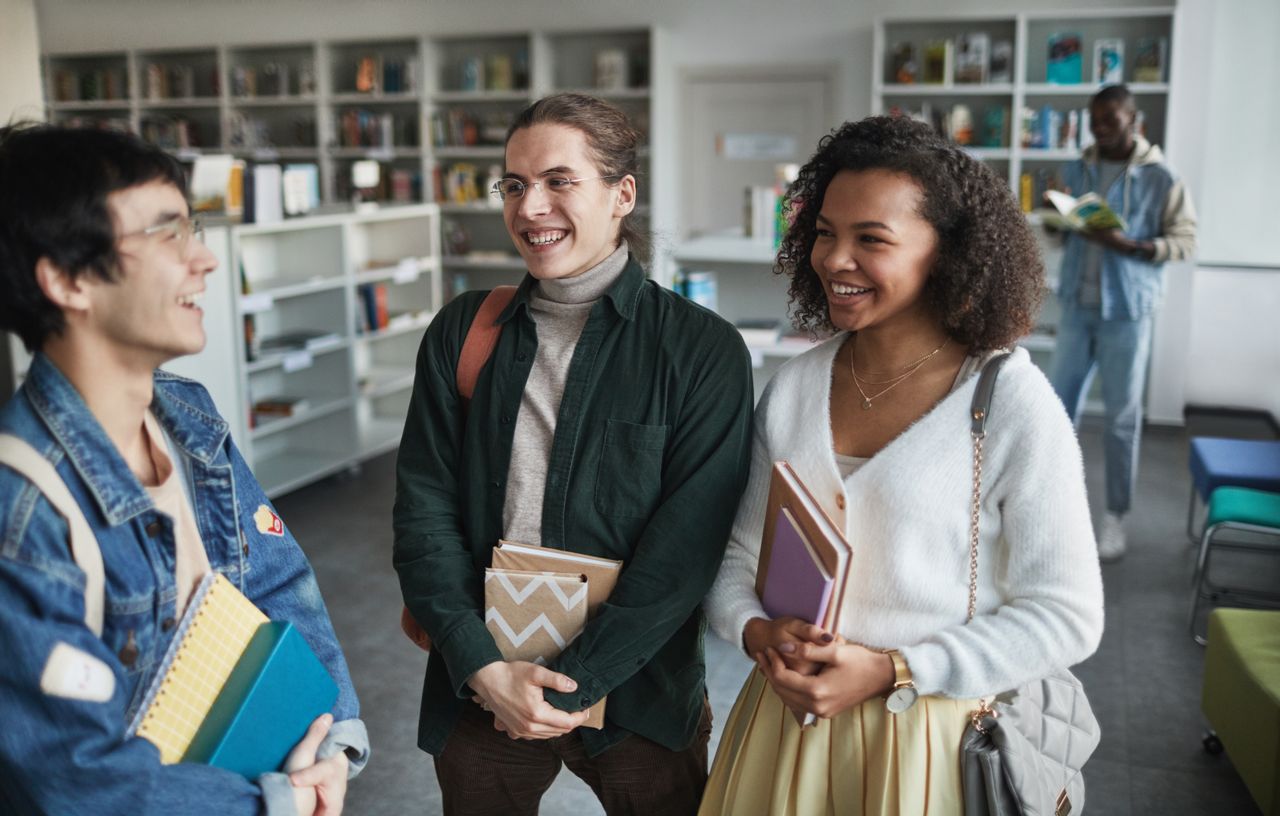 why-choose-us-01 Group of diverse students laughing and holding books in a library environment.