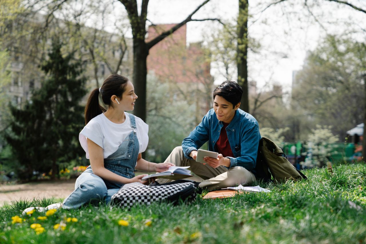 gallery-08 Two students studying together in a park on a sunny day, surrounded by nature.